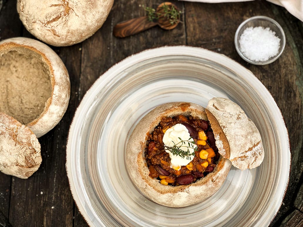 Herzhaftes Chili con Carne, angerichtet im ausgehöhlten Brottopf, serviert mit Sauerrahm und frischen Kräutern.
