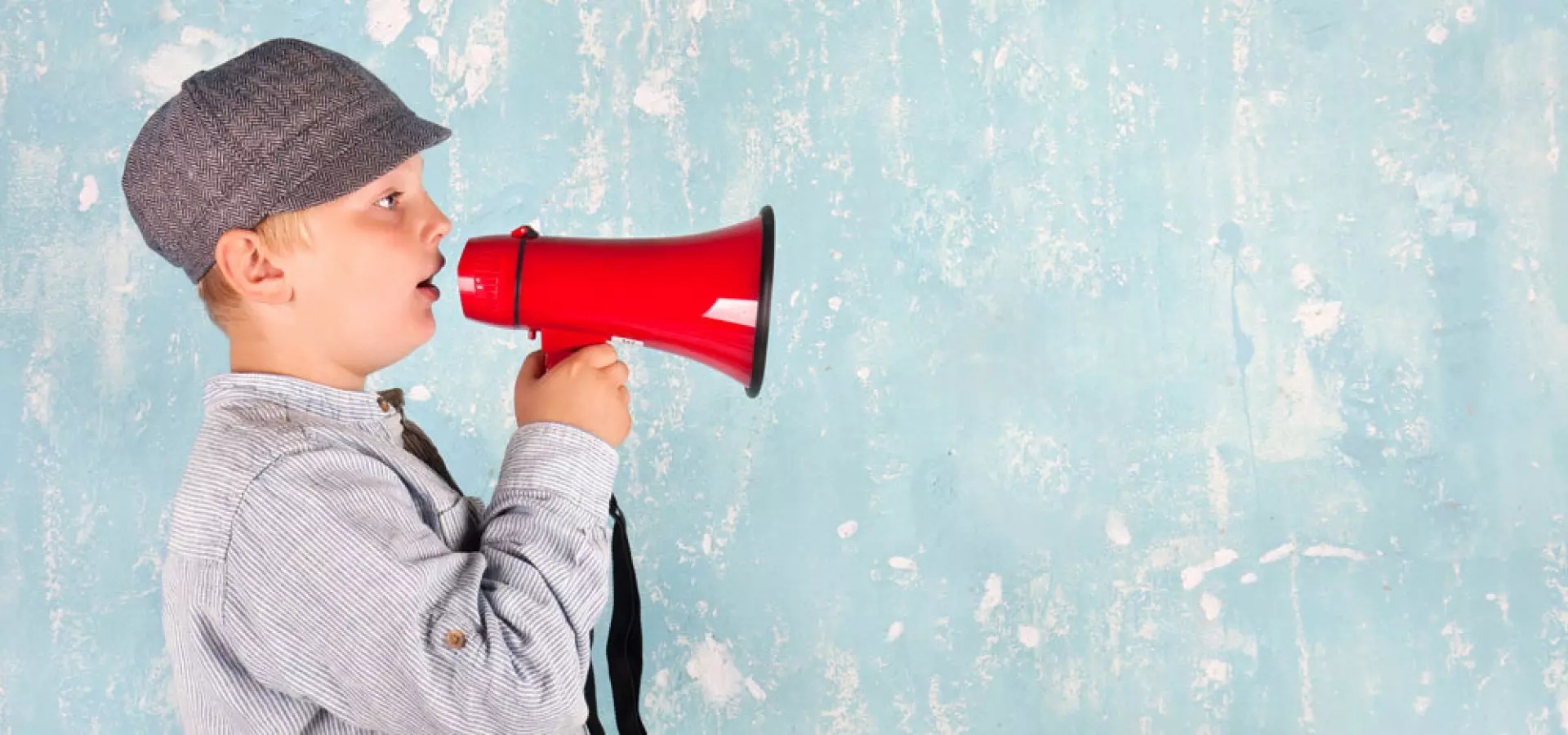 Kind, Megafon, Kommunikation, Ankündigung, Copyspace, Retro/Schiebermütze, aufmerksamkeitsstark. Möchtest du die Beschreibung als ganz kurzen Alt-Text (max. 125 Zeichen) oder für Social Media angepasst? Junge mit Schiebermütze hält ein rotes Megafon an den Mund und ruft nach rechts; Hintergrund ist eine hellblaue, abgeplatzte Wand mit viel Freiraum für Text